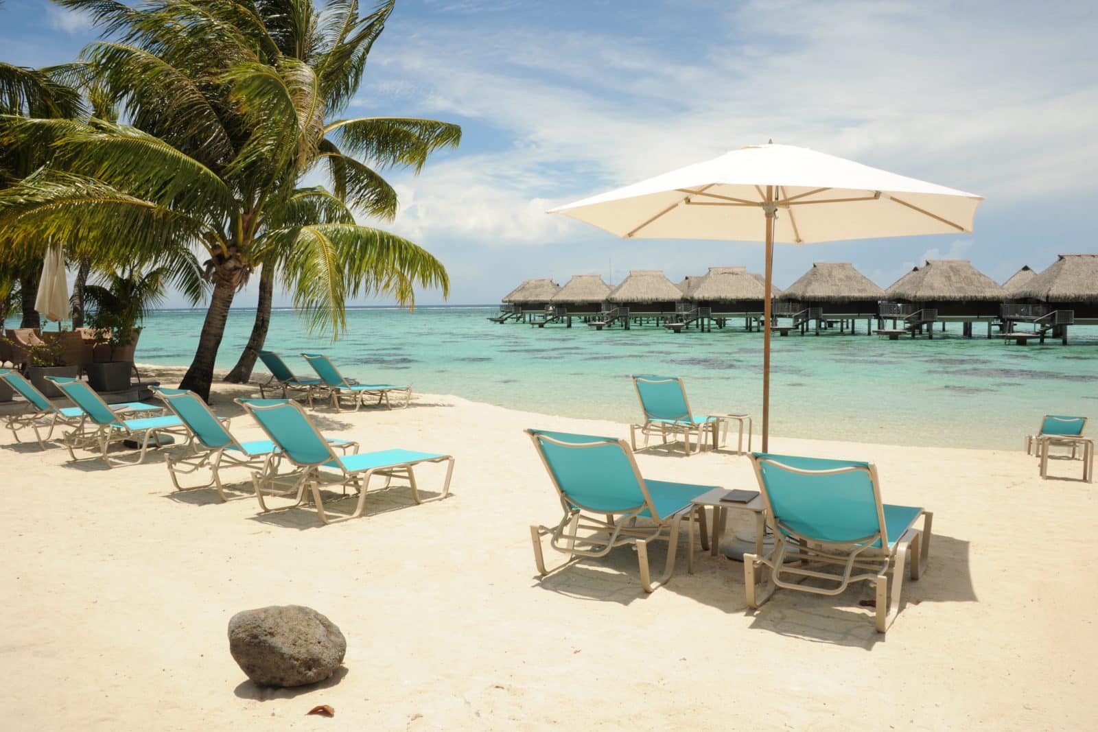 Plage à Morea, sable blanc, eau turquoise et transats sous l'ombre de palmiers. Au loin, des hébergements sur pilotis.